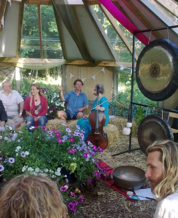 Group of people sitting in a greenhouse decorated with flowers and banners, with musical instruments like a cello and gongs.