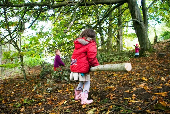 Children playing in a wooded area during autumn, with two girls in jackets. One girl in a red jacket and pink rain boots is walking uphill, holding a fallen tree branch, while another girl in a purple jacket is further up the hill, and a third child 