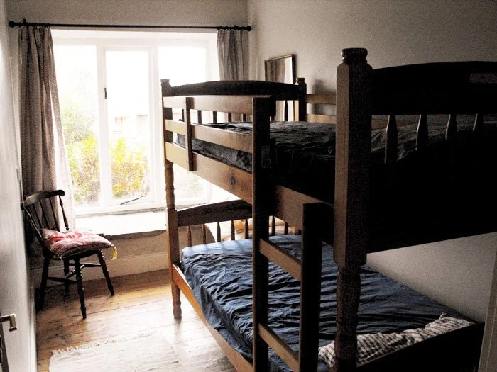 Room with a wooden bunk bed and a window with curtains, a chair with a cushion, and wooden flooring.