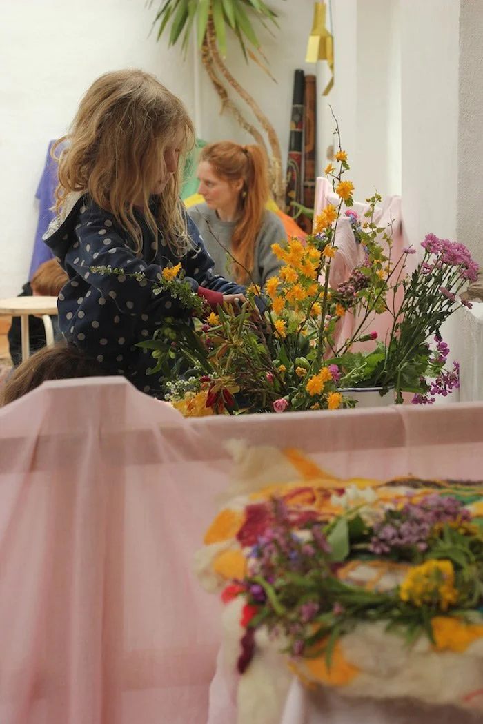 A young girl with blonde hair arranging flowers in a colorful bouquet, with a woman with red hair and a gray sweater observing in the background. There are additional flowers and greenery around them.