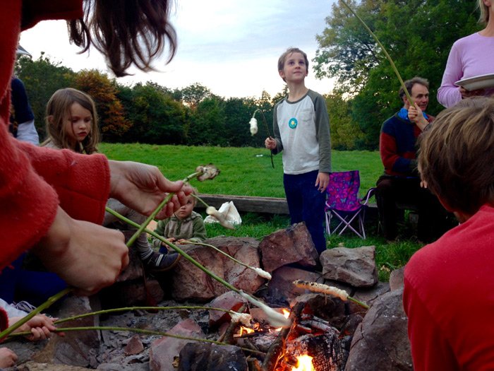 Children and adults roasting marshmallows over a campfire in a grassy outdoor setting.