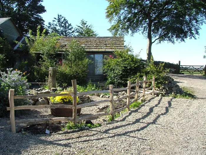 A rustic garden with a small wooden house, surrounded by plants and trees, enclosed by a simple wooden fence on a gravel path.