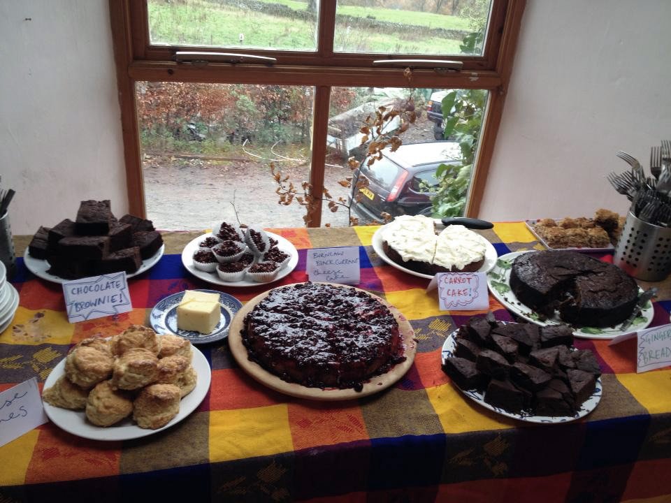 A table with various desserts including chocolate brownies, cupcakes, carrot cake, and assorted slices of chocolate and fruit cake, set in a room with a window overlooking a garden.