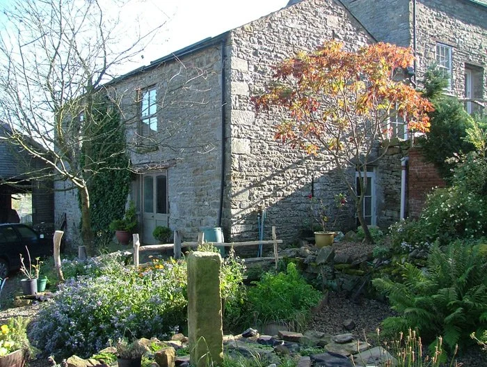 A stone house with a garden in front, featuring trees, plants, and flowers.