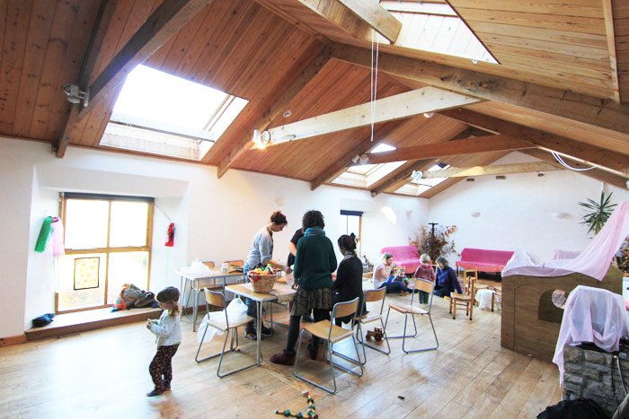 People gathered in a spacious room with a wooden ceiling, skylights, and large windows, engaged in activities like making crafts or preparing food, with children present in a cozy setting.