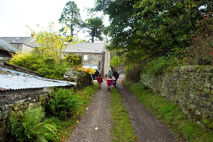People walking up a dirt path lined with greenery and stone walls, with houses and trees in the background.