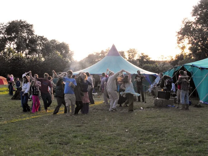 People dancing and enjoying live music outdoors near a large blue and green carnival tent at sunset.