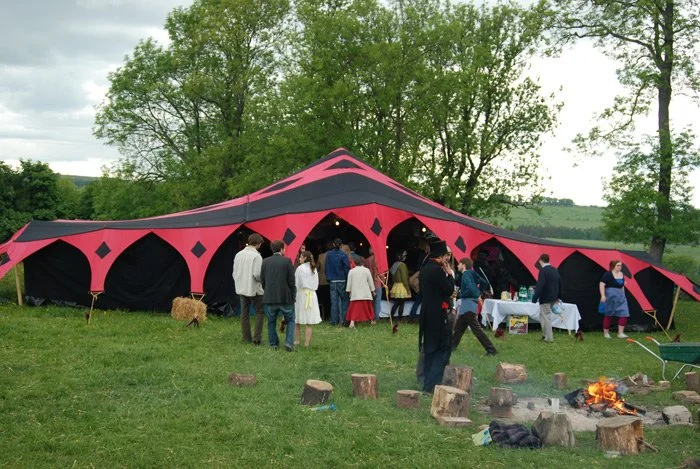 People gathering outside a large red and black tent in a grassy field with trees and a firepit.