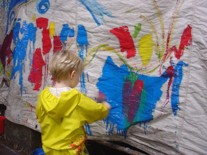 Young child in a yellow smock painting abstract colors and shapes on a large white fabric with vibrant blue, red, yellow, and green paint.