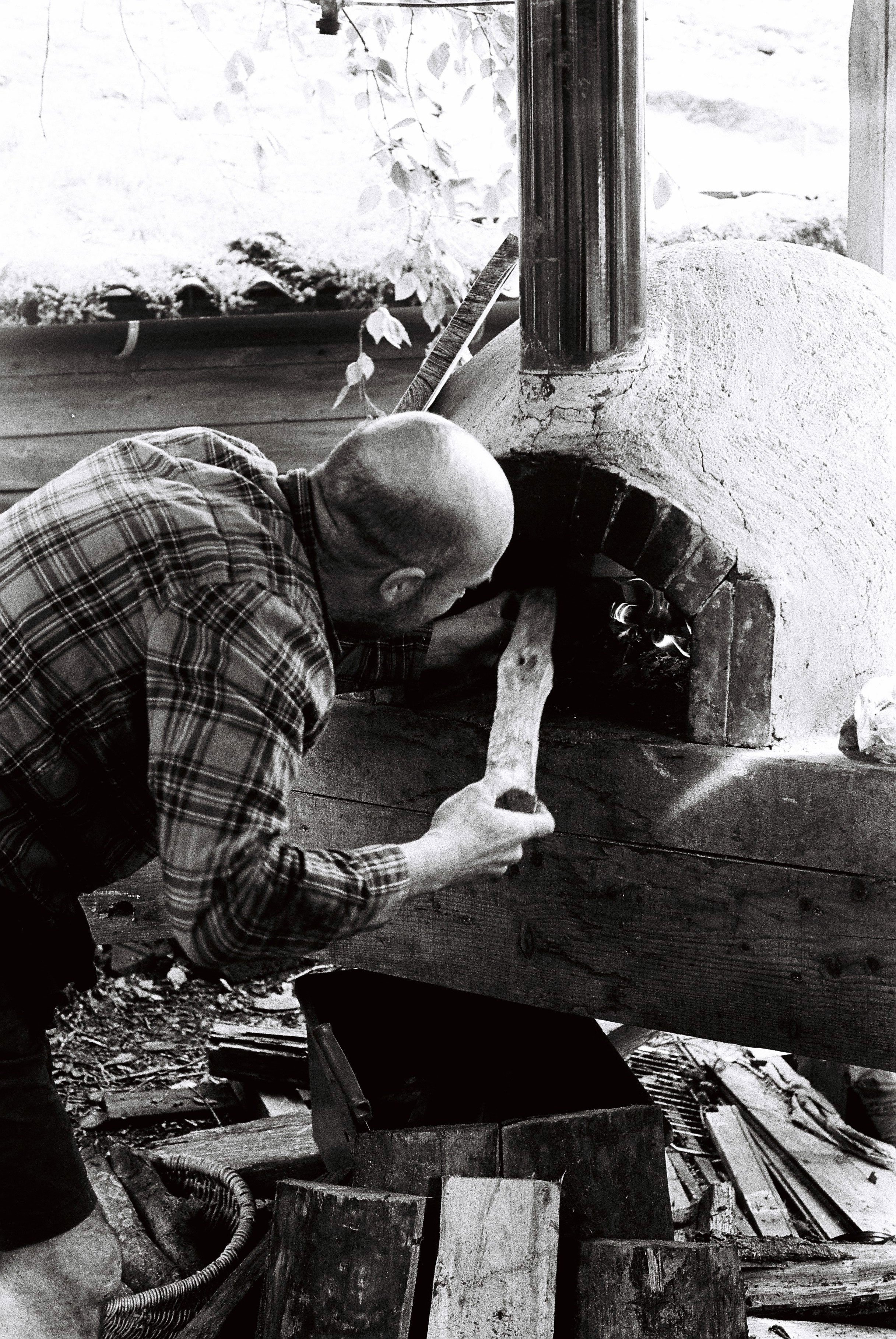 A man in a plaid shirt working on a brick oven, using a hammer to make adjustments.