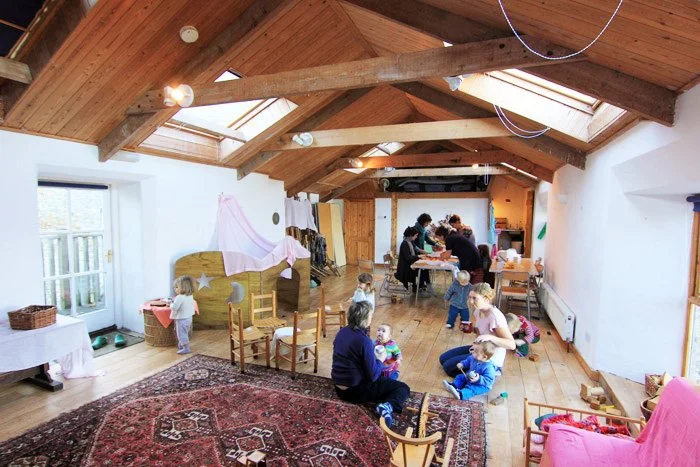 Group of children and adults in a spacious attic room with a wooden ceiling, skylights, and hardwood floors, engaged in various activities such as playing and crafts.