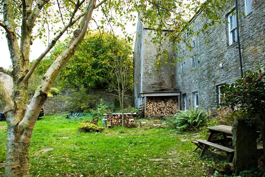 Backyard with green grass, trees, a wooden bench, and a stone building with a woodpile under an overhang.