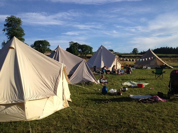 Tent tents set up on grassy field with people and camping equipment, blue sky with clouds in the background.