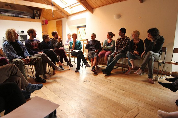 People sitting in a semi-circle in a cozy, well-lit room with wooden floors and walls, engaging in a group discussion.