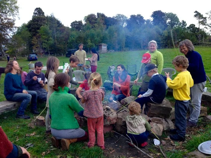 Group of children and adults around a campfire outdoors, some sitting on logs and others standing, with trees and a grassy field in the background, enjoying a social gathering.