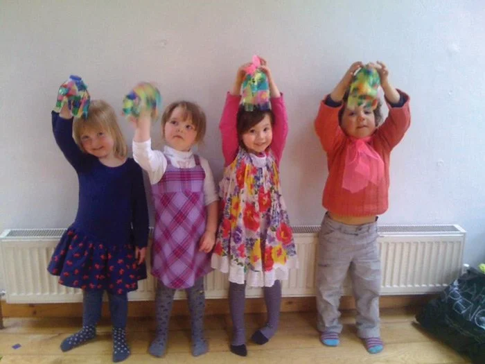Four young girls standing indoors, holding colorful pom-poms above their heads.