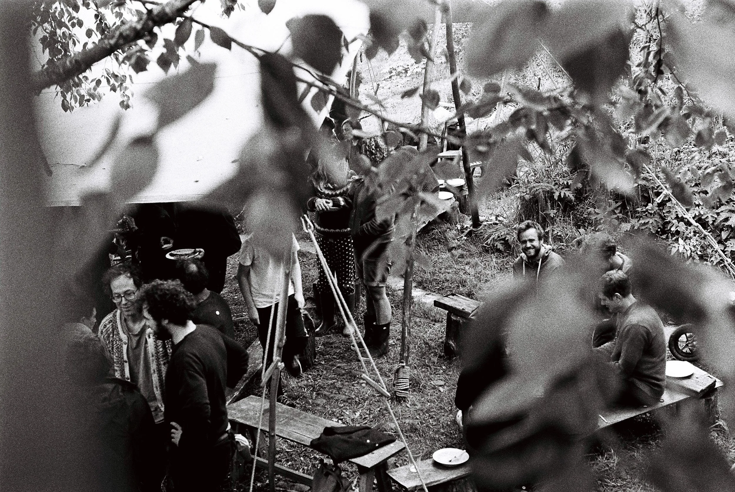 A group of people gathered outdoors, some sitting and some standing, surrounded by trees and leaves. The scene appears to be casual socializing in a natural setting.