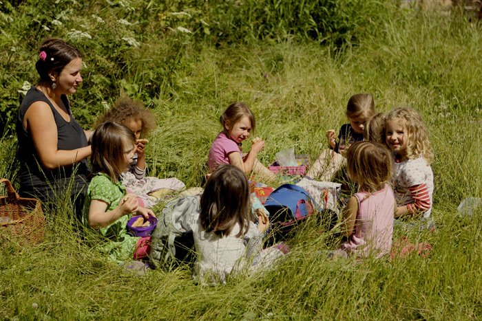 Group of young girls having a picnic outdoors in a grassy field, sitting around a blanket with snacks and drinks, with an adult woman supervising.