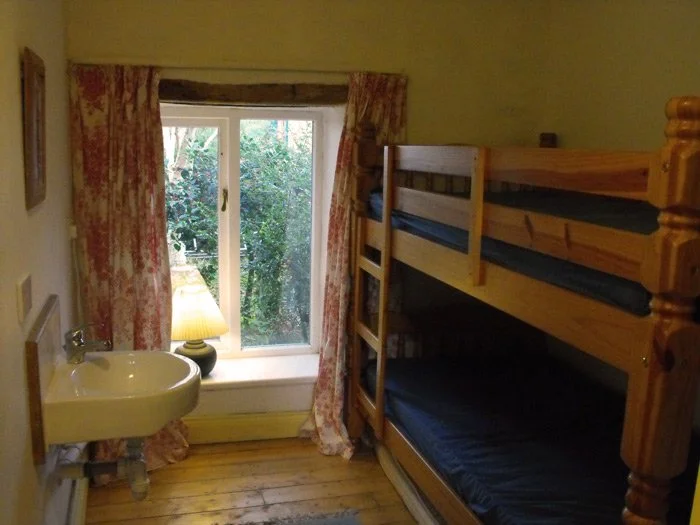 A small bedroom with a wooden bunk bed, a window with floral curtains revealing greenery outside, a small sink with a lamp on the windowsill, and wooden flooring.