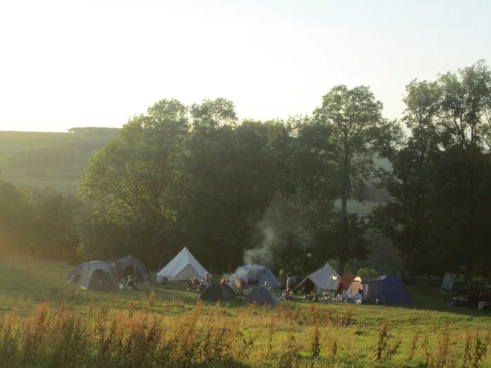Tents and camping setup in a grassy field with trees in the background, early morning or late afternoon sunlight.