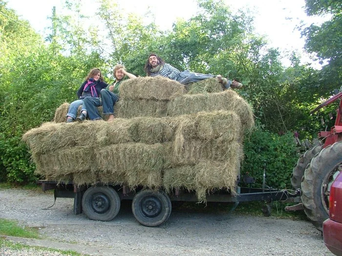 Three people sitting and lying on large hay bales stacked on a flatbed trailer, attached to a tractor, outdoors in a rural area with trees in the background.