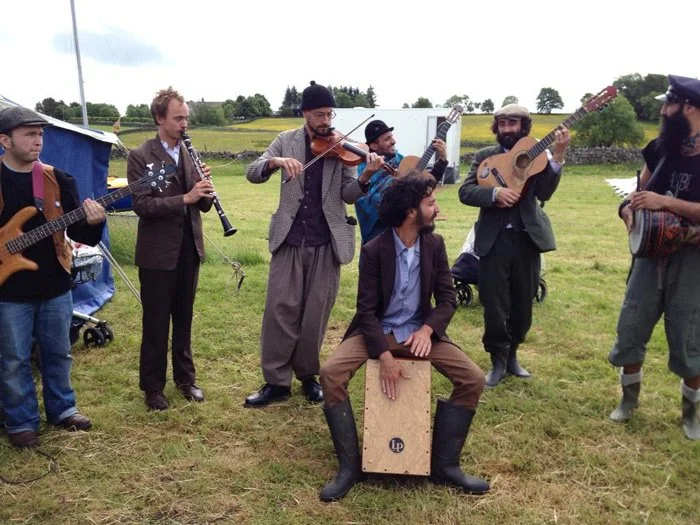 Group of musicians playing instruments outdoors on a grassy field, including guitars, a violin, a clarinet, and a cajón, with some musicians sitting and others standing.