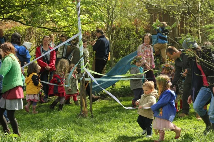 Children and adults participating in an outdoor spring festival or event, with some children playing near a decorated area with ribbons and trees.
