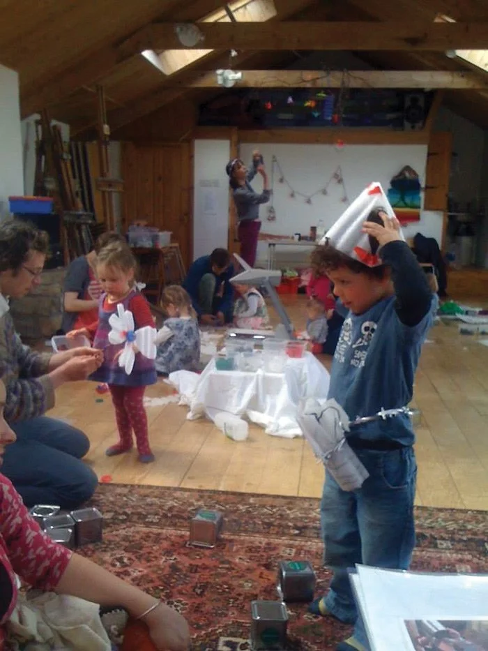 Children and adults participating in a holiday craft workshop in a cozy wooden attic with a sloped ceiling, creating paper angel decorations and wearing festive accessories.