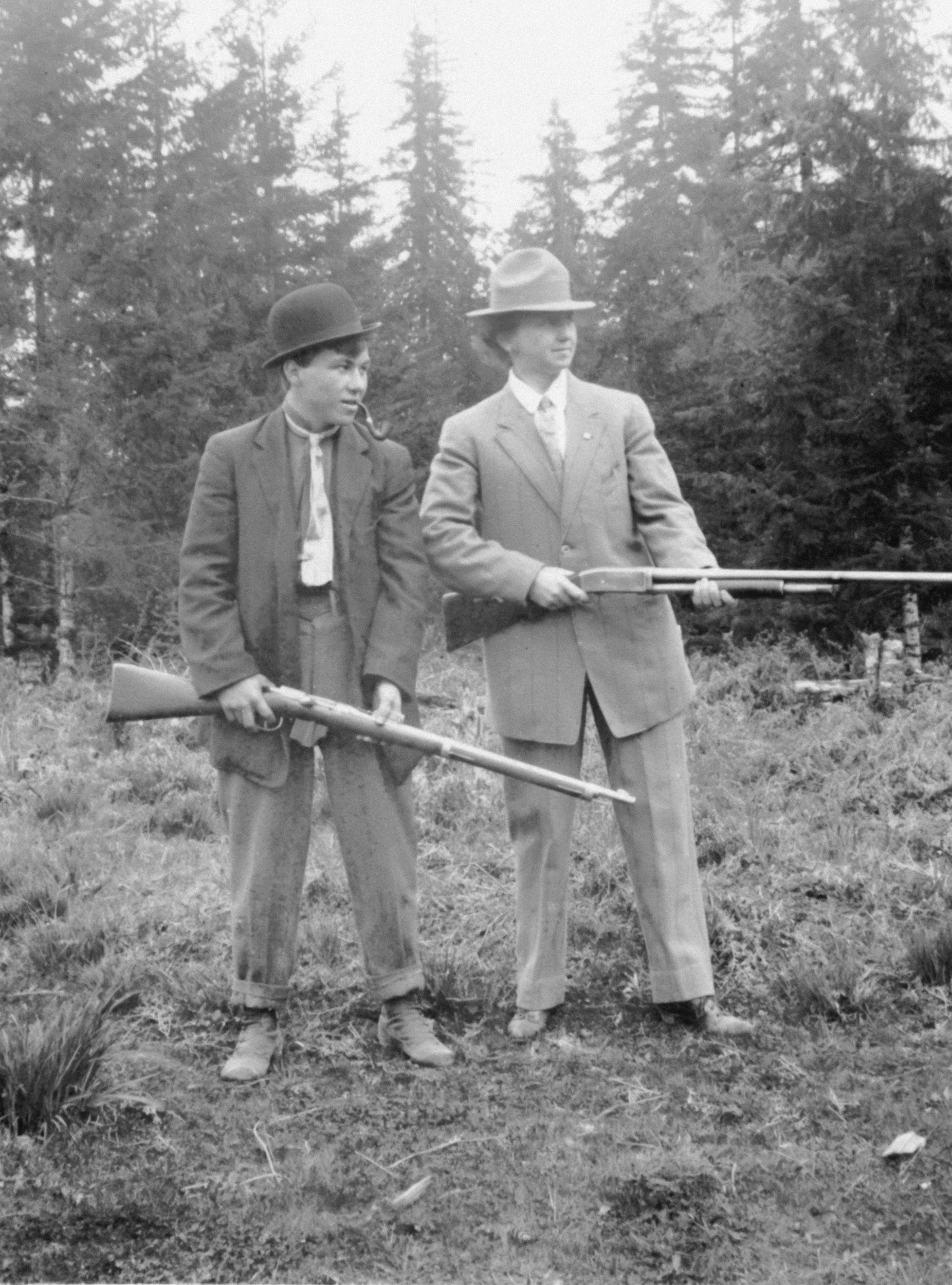 Two women in vintage suits and hats standing outdoors in a wooded area, holding rifles.