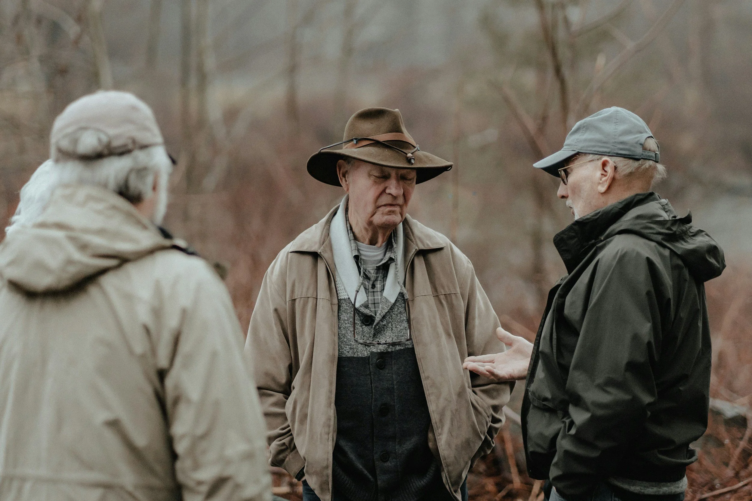 Four older men wearing outdoor jackets and hats, engaged in a conversation outdoors in a wooded area during fall.
