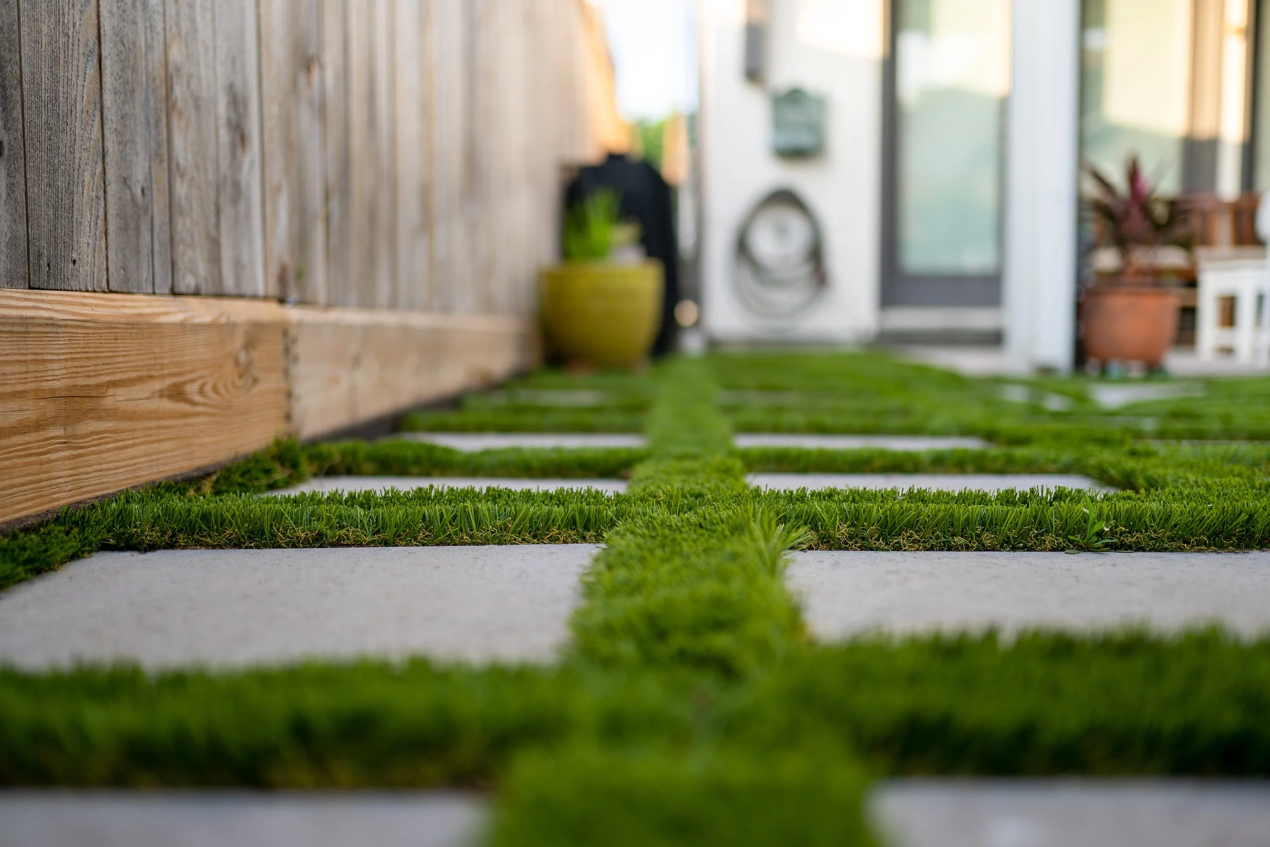 Close-up of a pathway with square concrete slabs and green grass between them, leading towards a house in the background.
