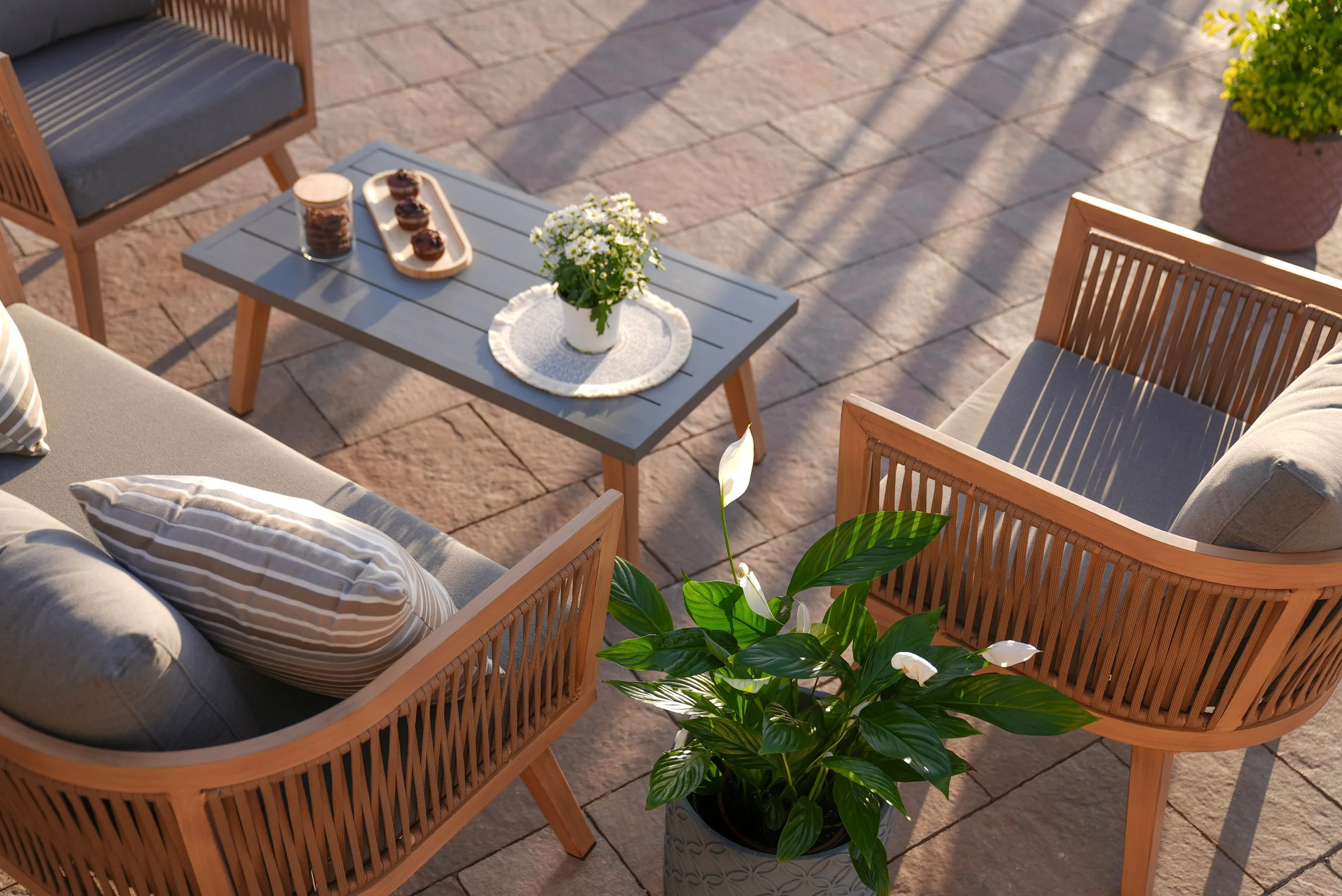 Outdoor patio with wooden furniture, including a cushioned sofa, two chairs, a gray coffee table with a vase of white flowers and a tray of chocolates, and a potted plant with white flowers. Sunlight creates shadows on the paved surface.