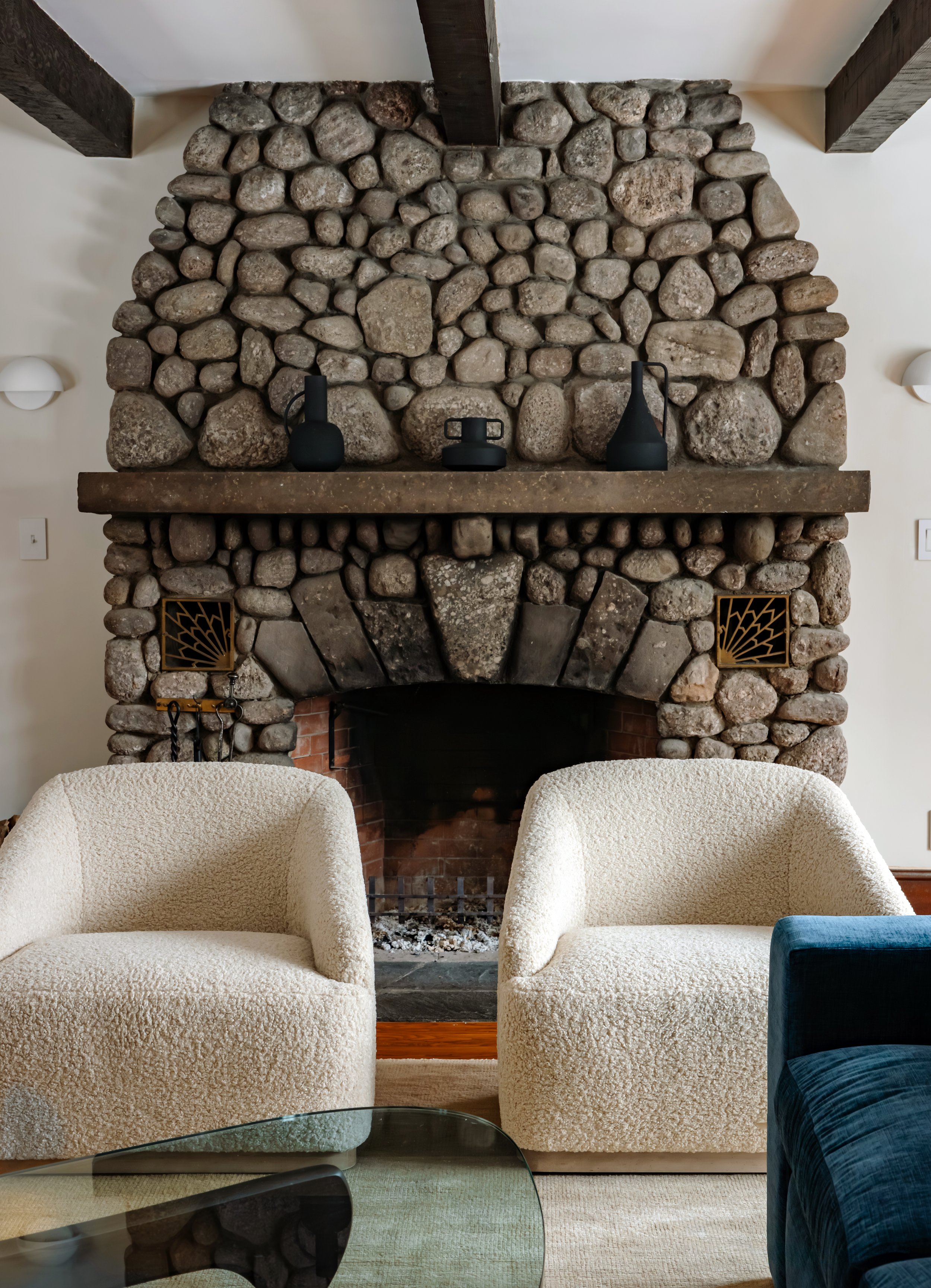 Rustic stone fireplace with sculptural black decor and two boucle lounge chairs.