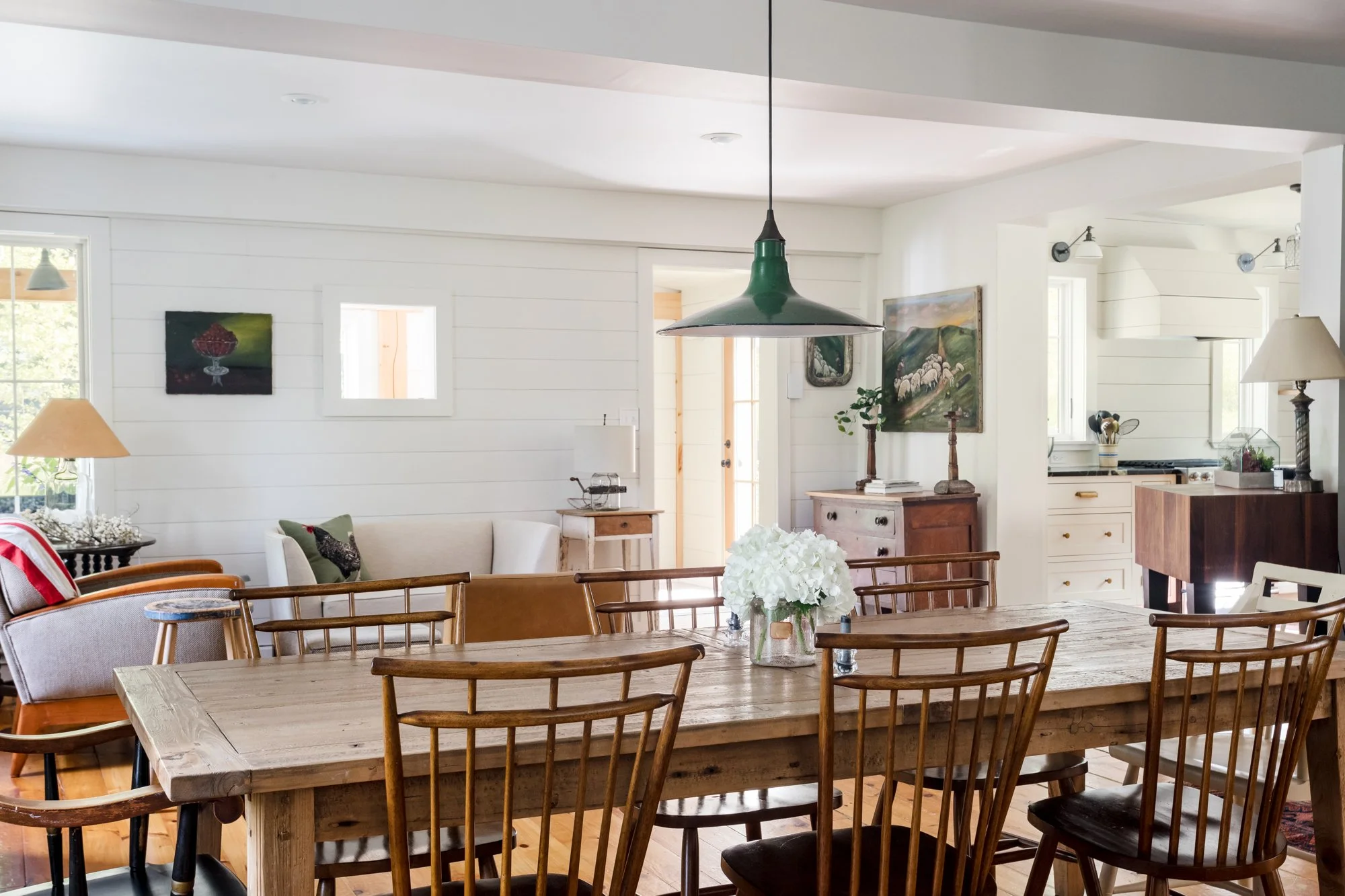 A cozy, bright dining and living room with wooden furniture, white walls, and natural light coming through windows. Decor includes paintings, table lamps, and a vase with white flowers on the dining table.