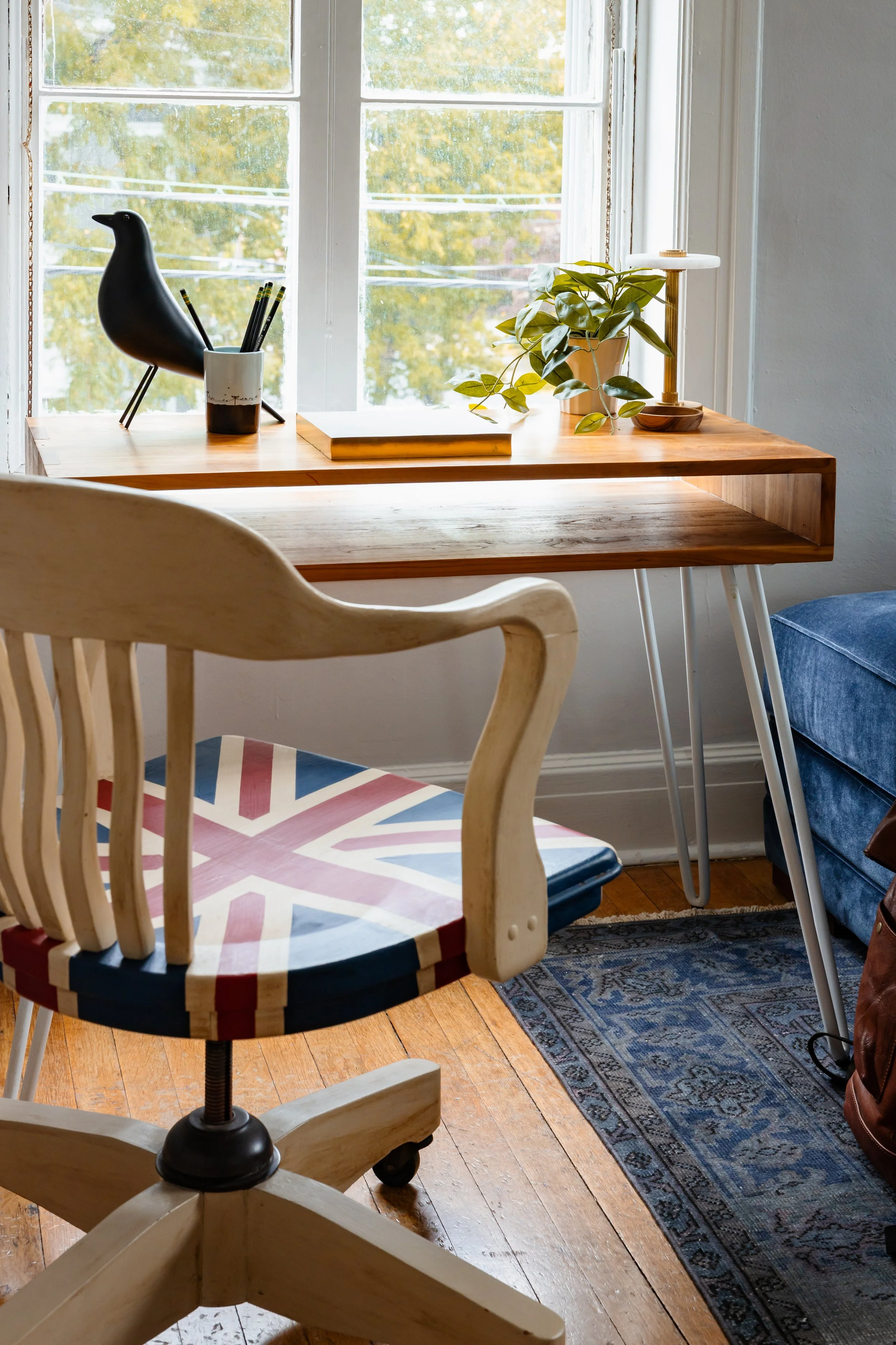 Small wood desk styled by a tall window with plant, lamp, and vintage office chair.