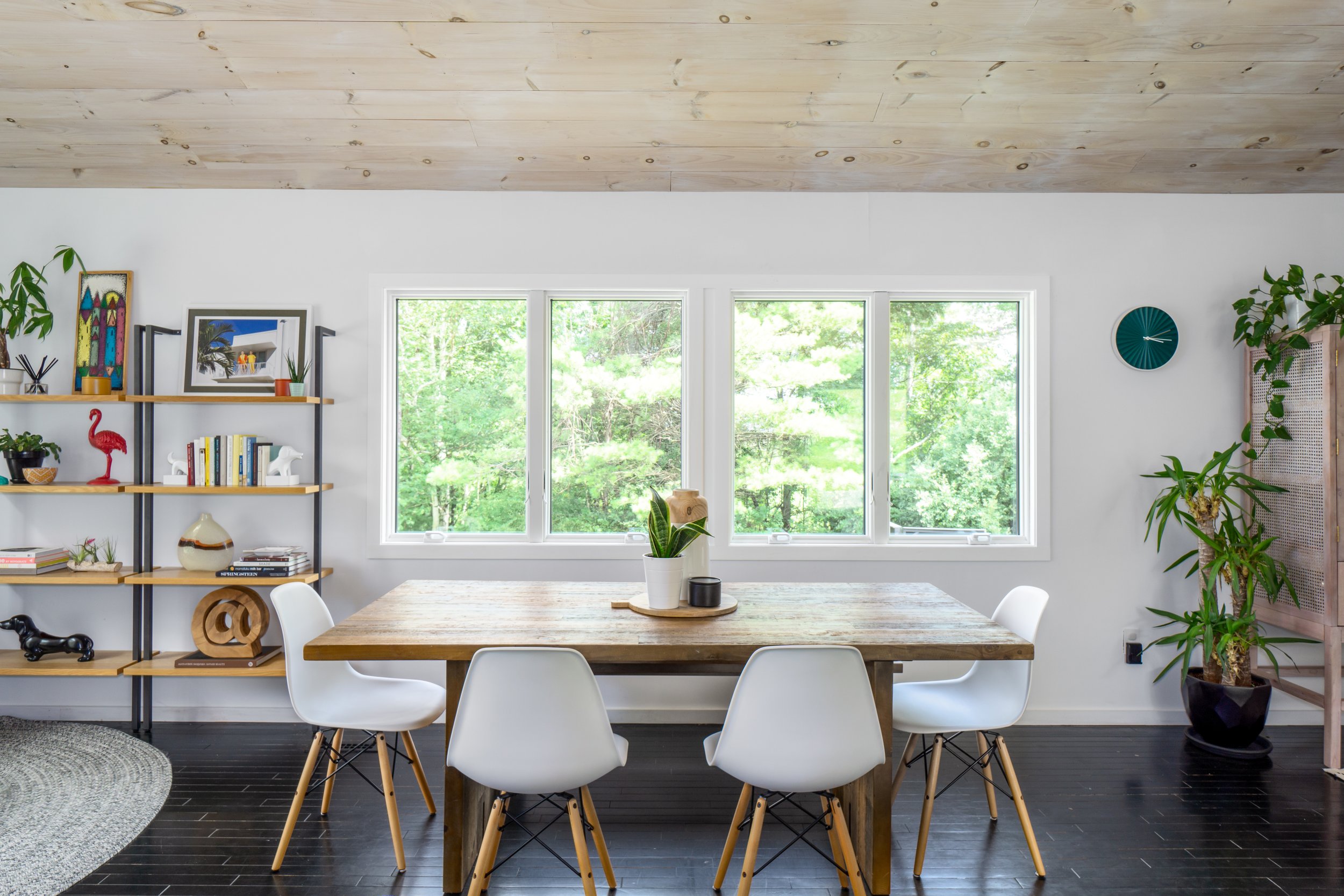 Dining room with rustic wood table, white chairs, and large windows overlooking trees.