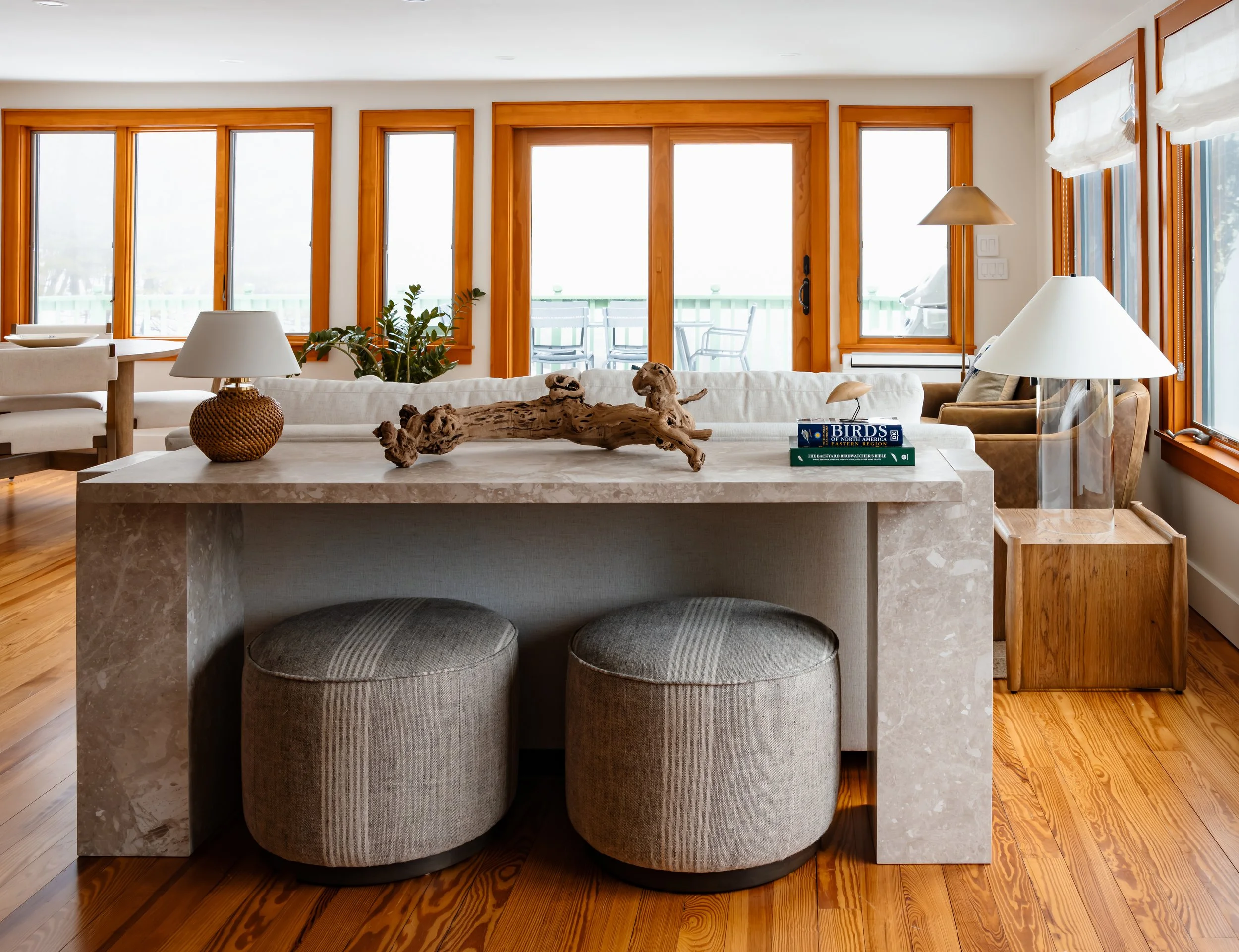 Stone console table with upholstered ottomans in a bright sitting area with wood-trimmed windows.