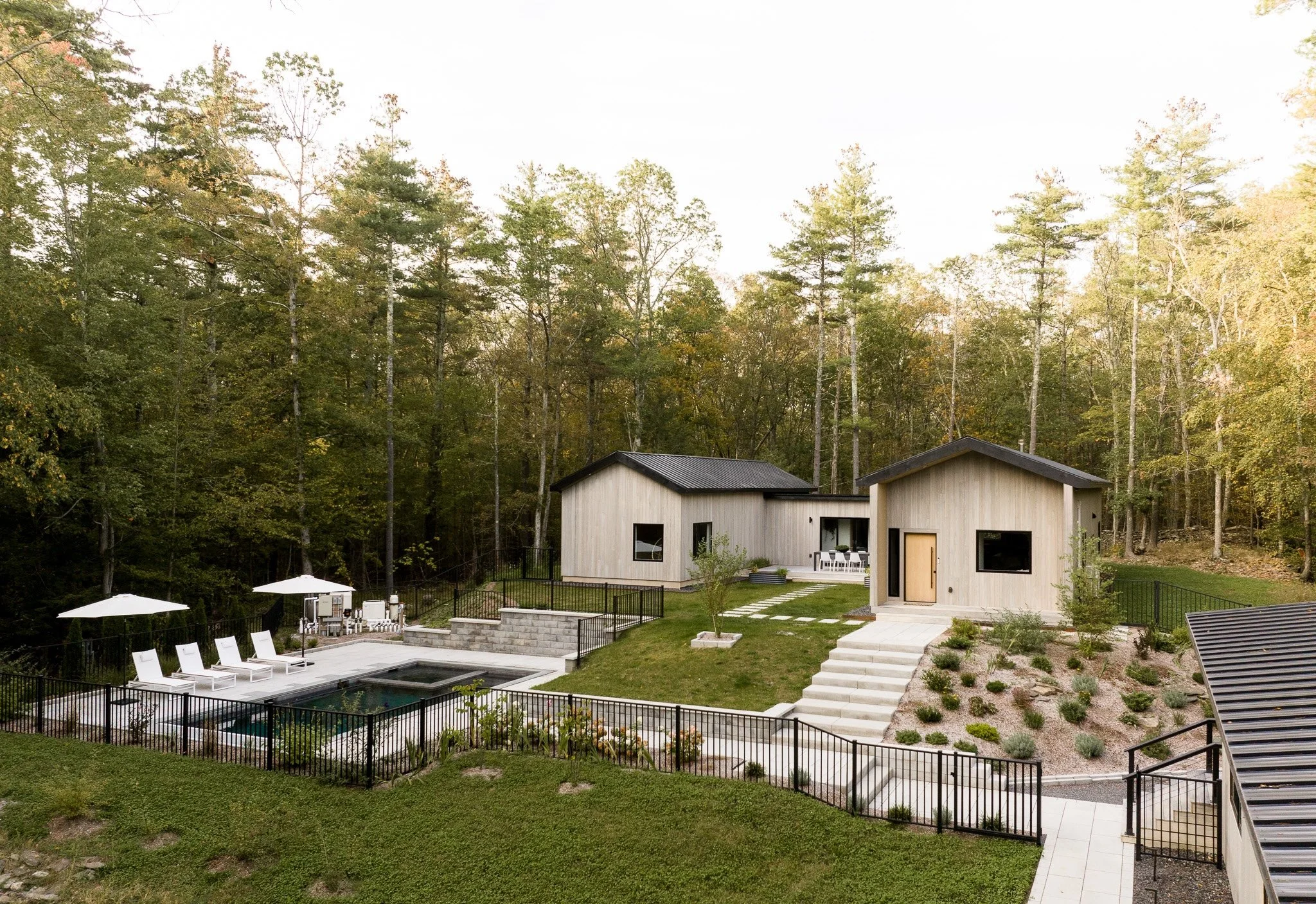 Modern wood-clad home with two gabled structures, landscaped yard, and in-ground pool surrounded by forest.