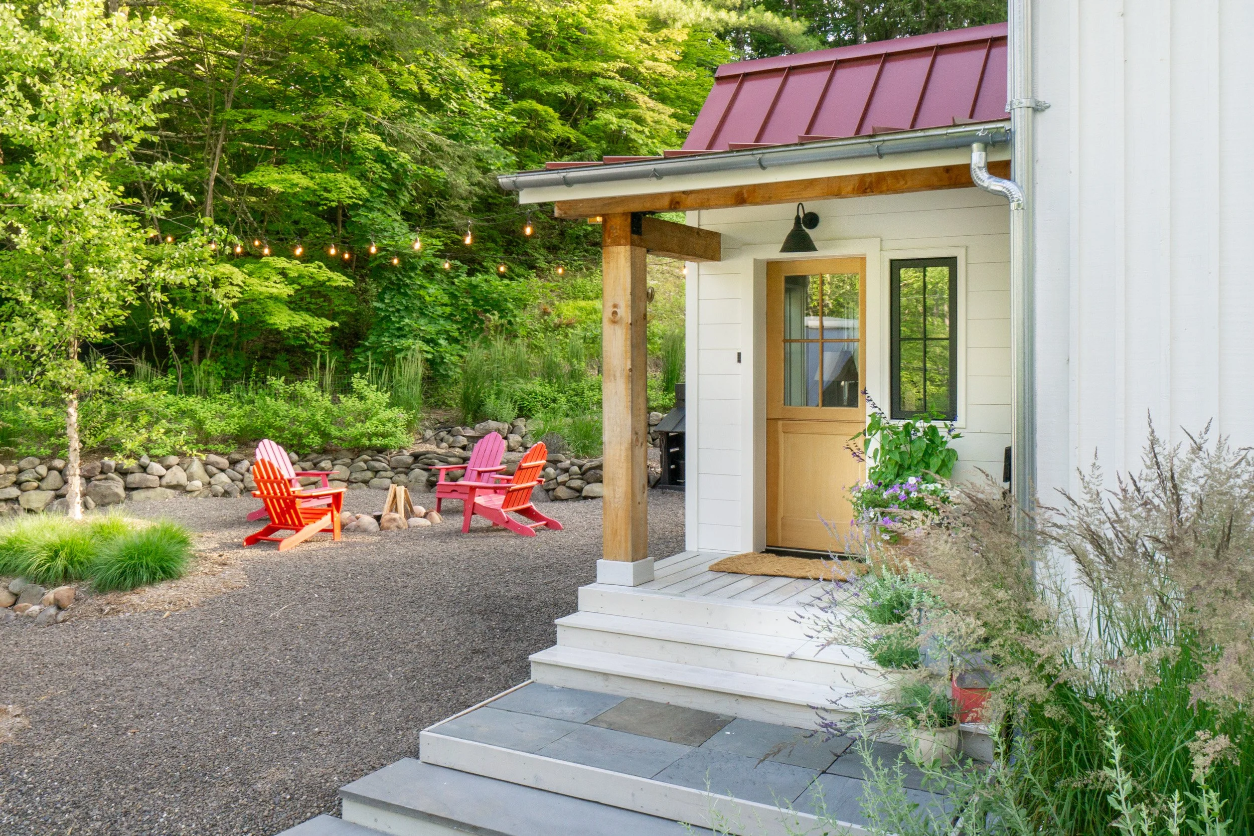 Front porch of white cottage with red metal roof and firepit seating area in landscaped yard.