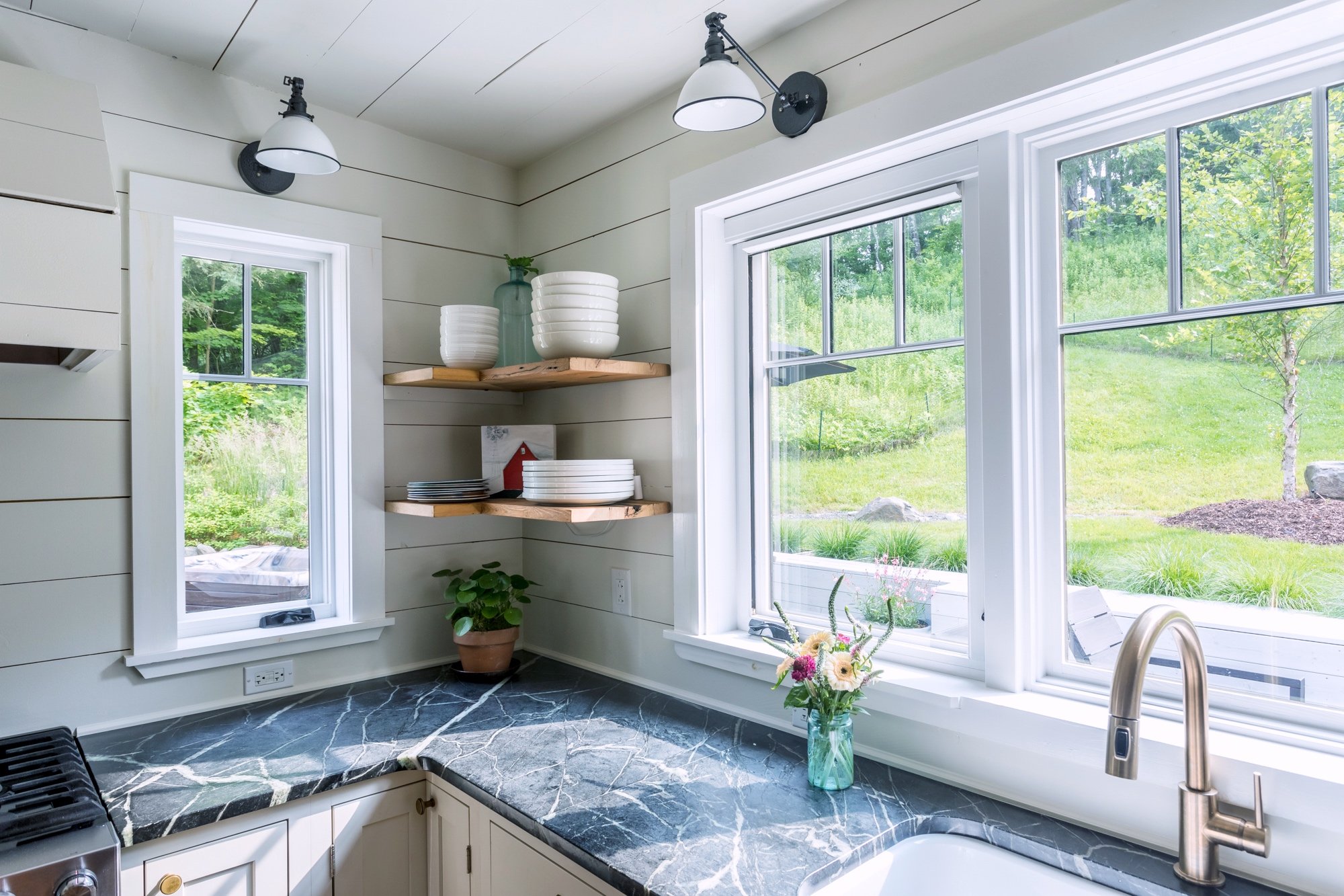 Kitchen corner with open wood shelving, white shiplap walls, and large windows overlooking greenery.