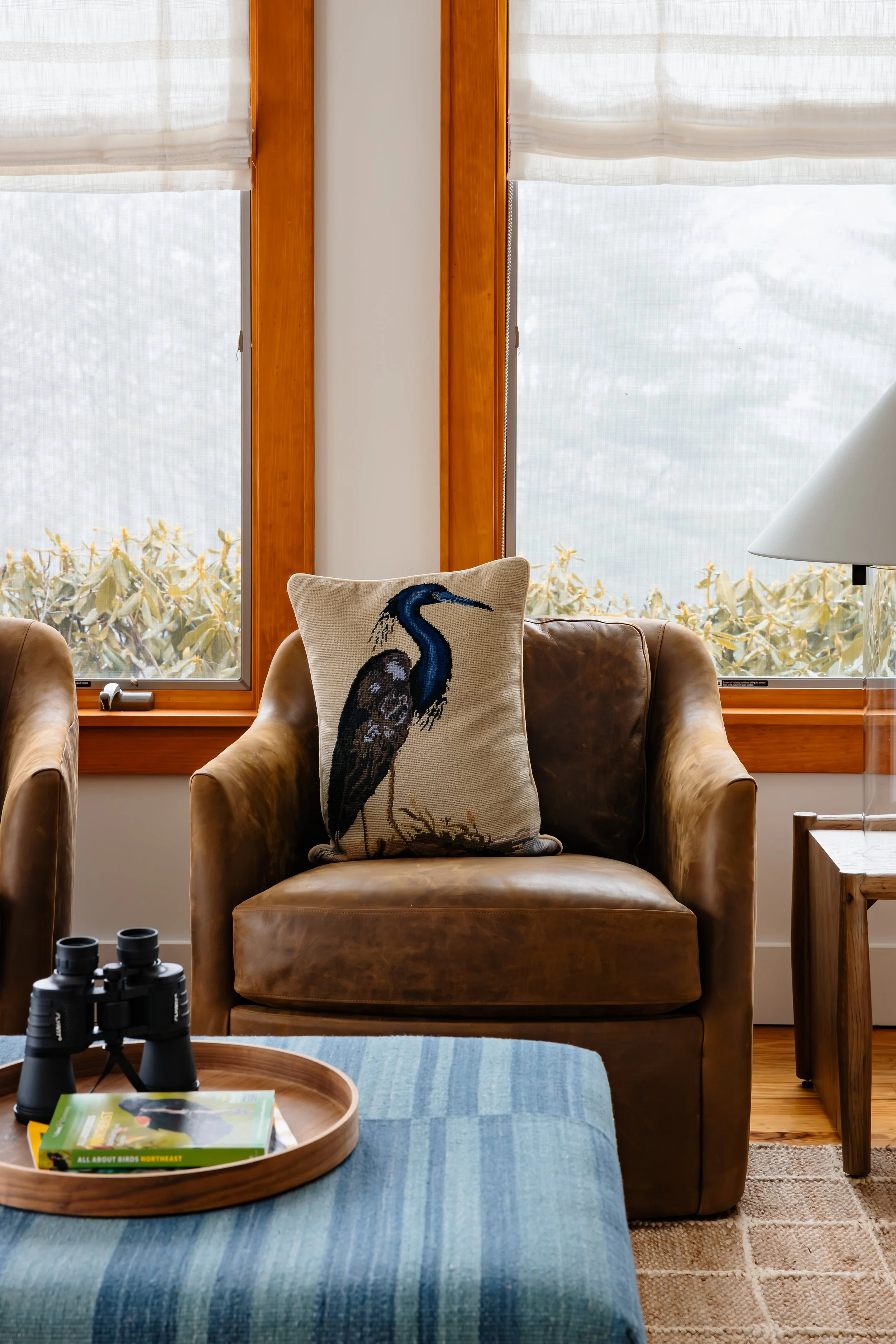 Leather lounge chair with bird motif pillow in front of wood-trimmed windows.