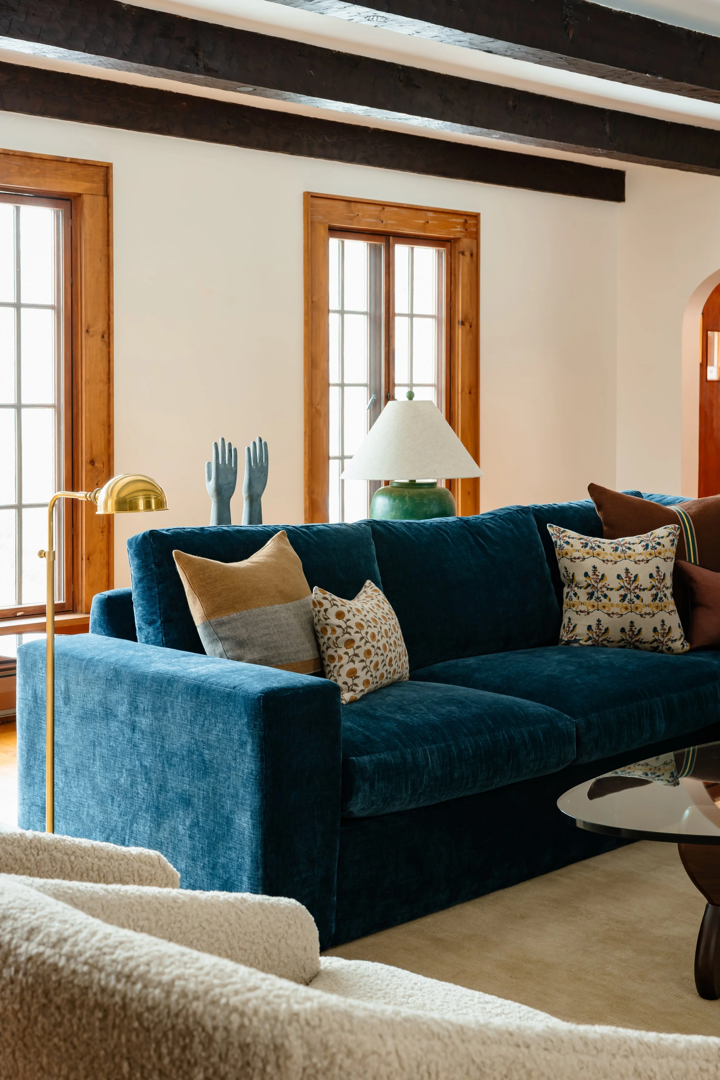 Blue velvet sofa with layered pillows set beneath dark wood ceiling beams and tall windows.