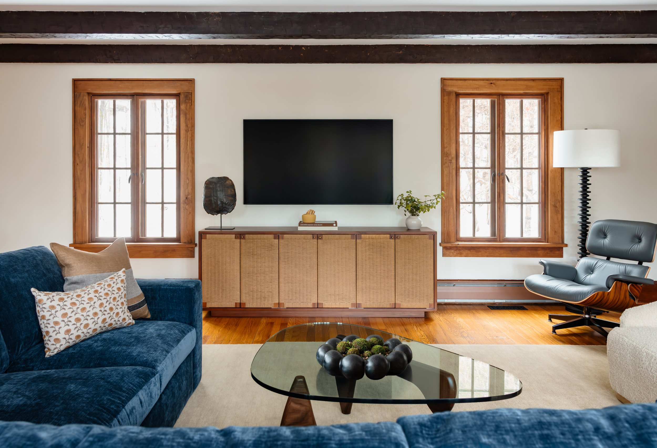 Living room with blue sectional, wood-framed windows, woven media cabinet, and glass coffee table.