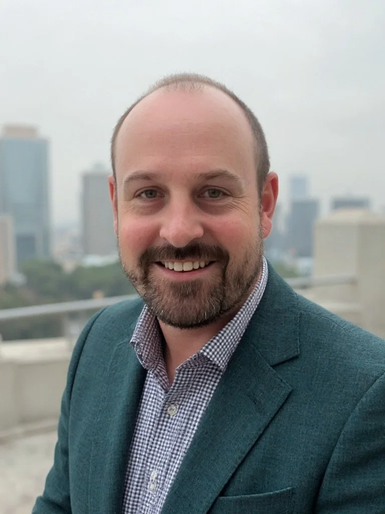 A smiling man with a beard and receding hairline, wearing a teal blazer and a checkered shirt, standing outdoors with a city skyline in the background.