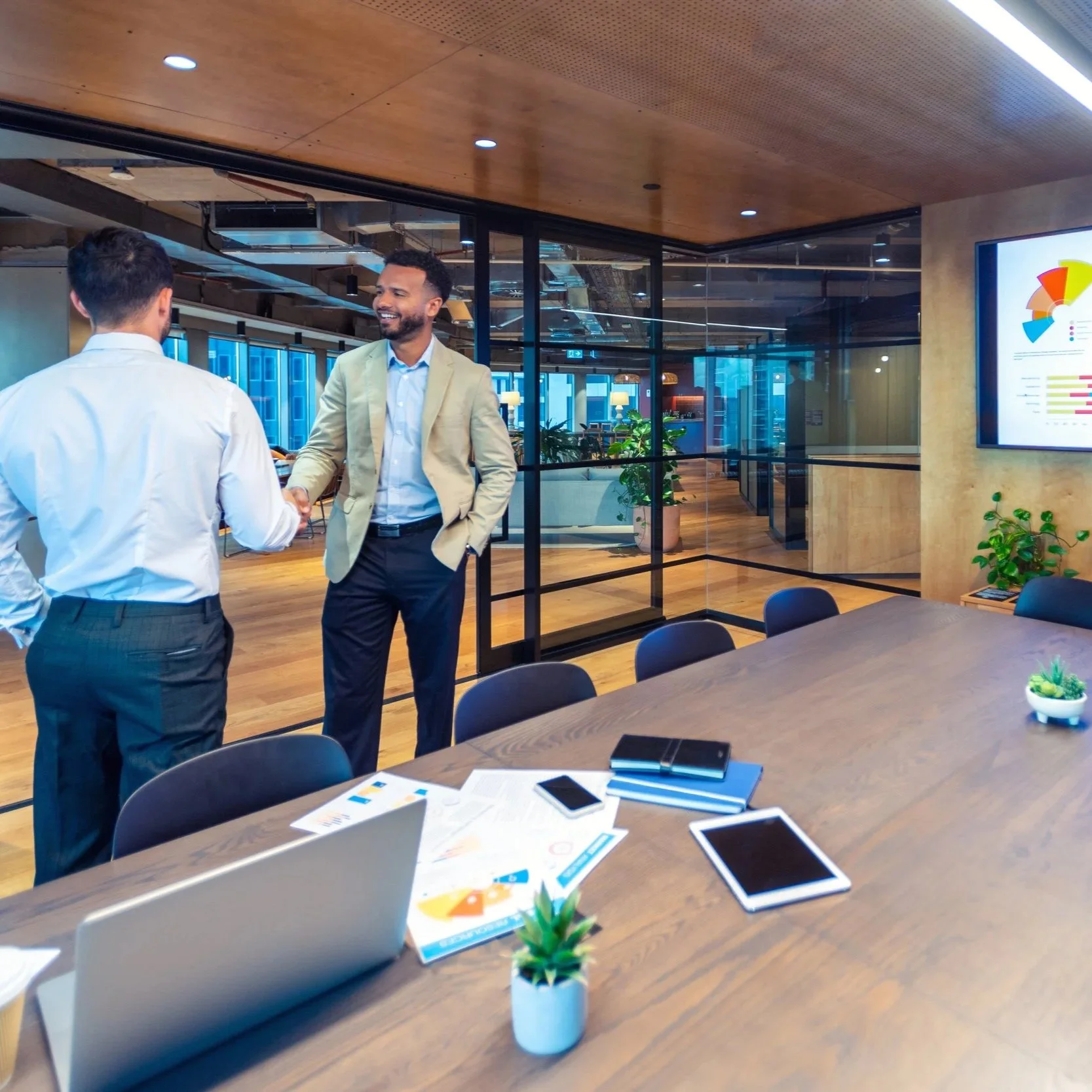 Two businessmen shaking hands in a modern office conference room, with a large table, laptops, tablets, documents, and plants on the table, and a digital chart on the wall.