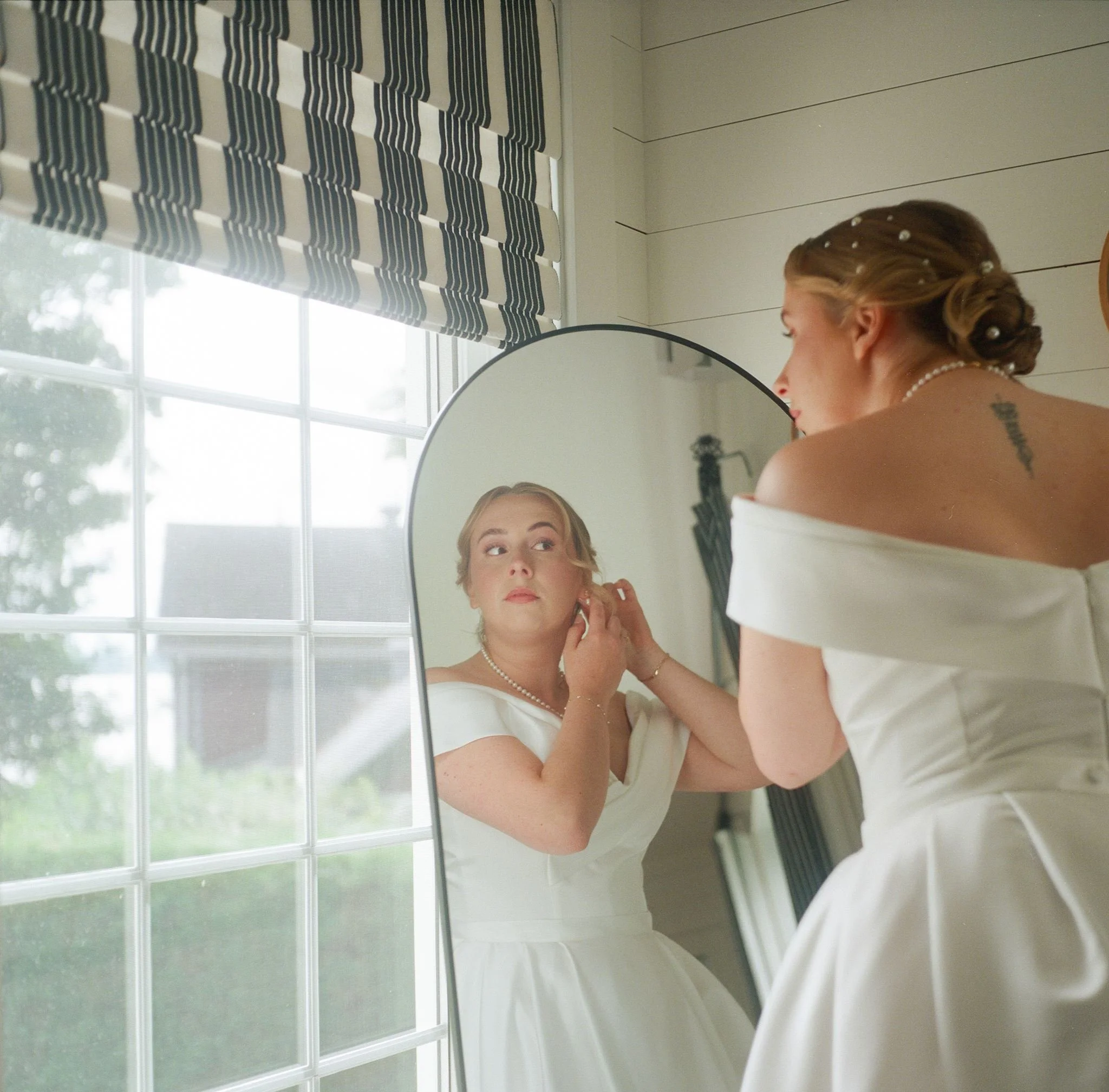 Une femme en robe blanche avec un collier de perles se regarde dans un miroir, près d'une fenêtre avec un rideau à rayures noires et blanches, dans une pièce lumineuse.