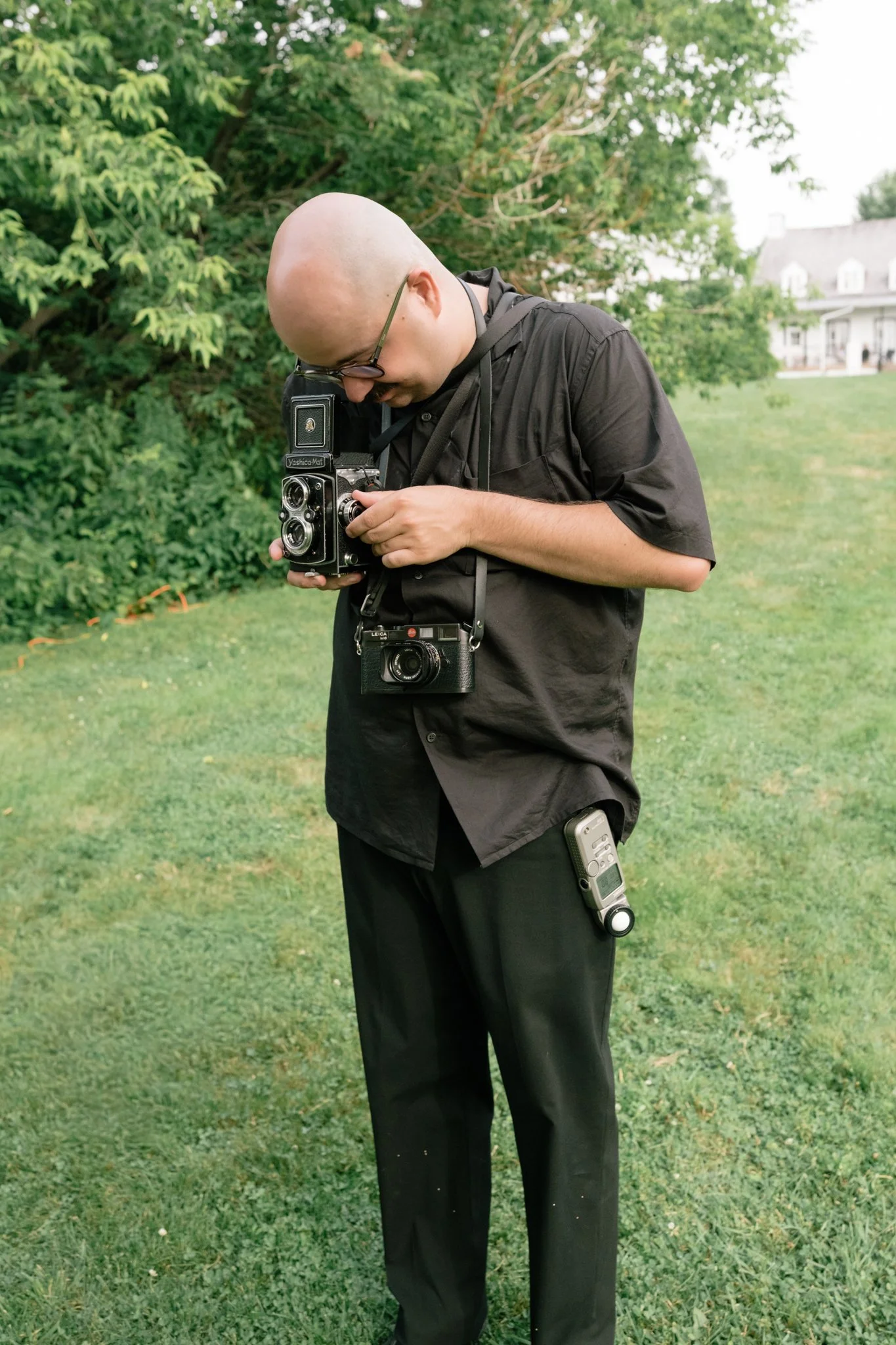 Homme portant des vêtements noirs, suspendu dans un parc verdoyant avec des arbres, utilisant un appareil photo vintage, avec un autre appareil accroché à la ceinture.