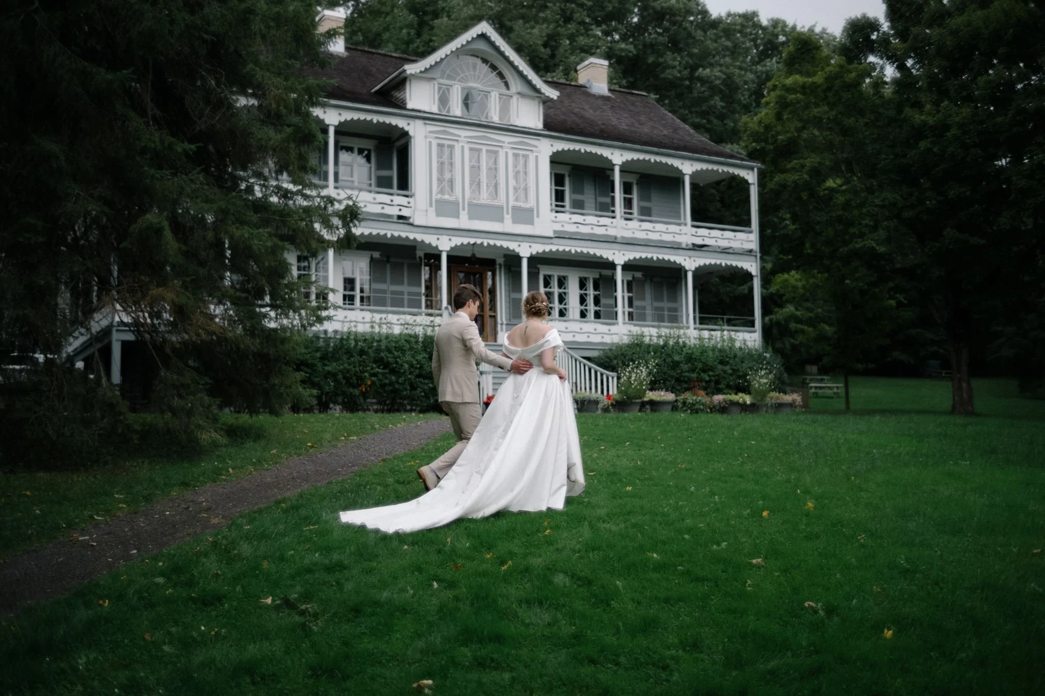 Un couple de mariés marche dans l'herbe devant une grande maison ancienne avec plusieurs porches, entourée d'arbres. La mariée porte une robe blanche longue, et l'homme un costume beige.