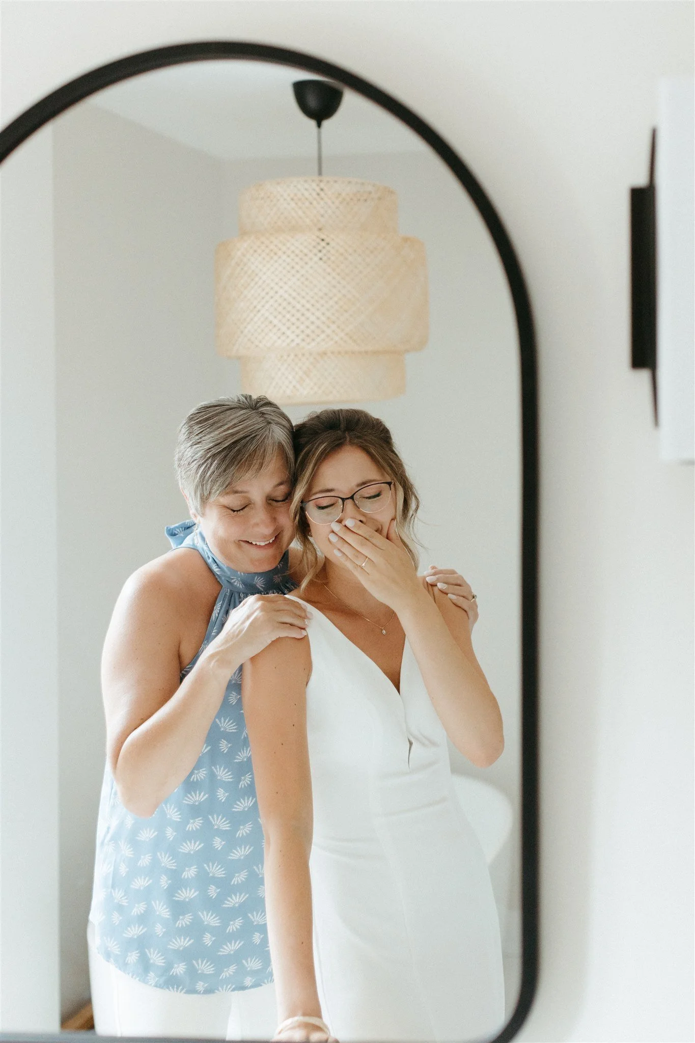 Deux femmes souriantes, l’une en robe blanche et l’autre en blouse bleue à motifs, se regardant dans un miroir et partageant un moment émouvant, captées par un Photographe mariage Québec à Québec.