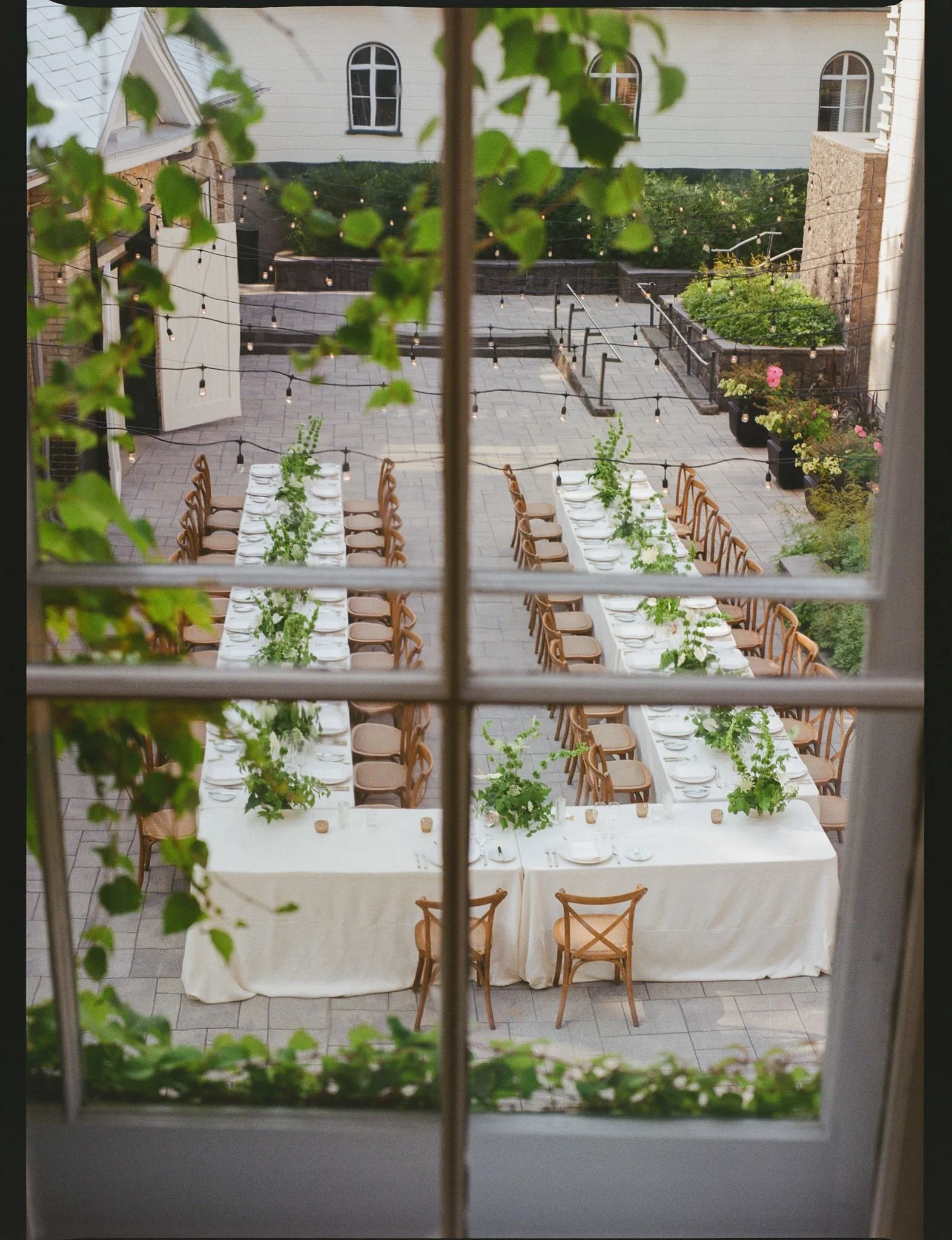 Vue d'une terrasse illuminée avec une longue table dressée pour un repas, des chaises en bois, des plantes vertes, des guirlandes lumineuses et une maison en arrière-plan vue de la fenêtre.